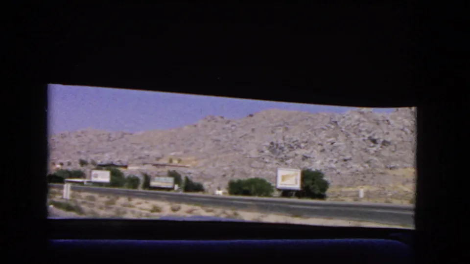 A 1966 California road-view frame showing billboard signs, shrubs, paved road, and rocky hillside in bright daylight.