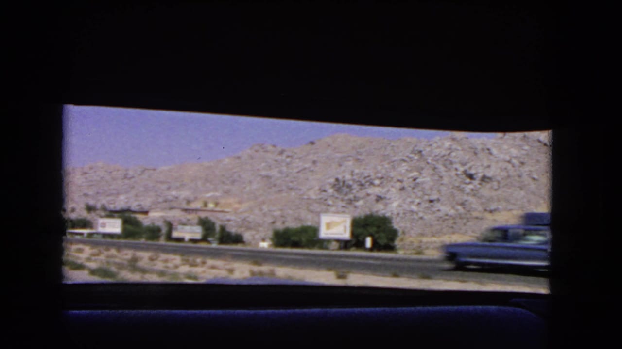 1966 California road view from inside a moving car, with rocky hills, roadside signs, sparse shrubs, and a blurred vintage car passing on the highway.