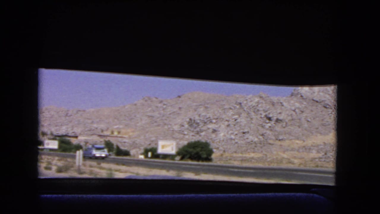 1966 California highway scene from a passenger window, showing an oncoming vintage car, roadside signs, scrub greenery, and rocky hills beyond the road.