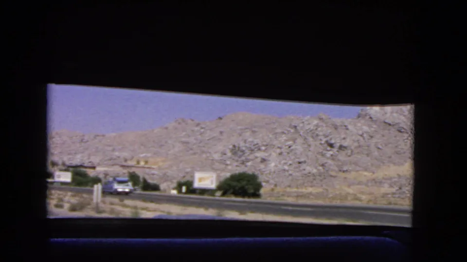 1966 California highway scene from a passenger window, showing an oncoming vintage car, roadside signs, scrub greenery, and rocky hills beyond the road.