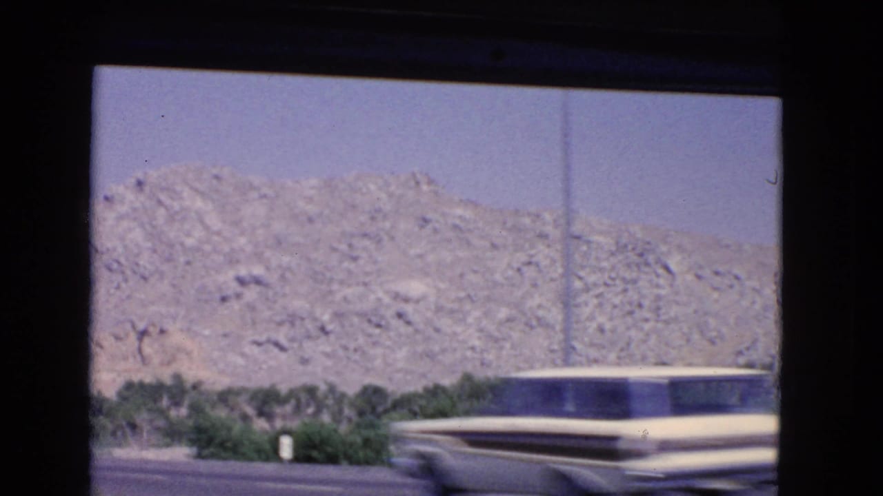 A blurred mid-century car passes close to the moving camera in a 1966 California roadside view with rocky hills, greenery, and a tall pole.