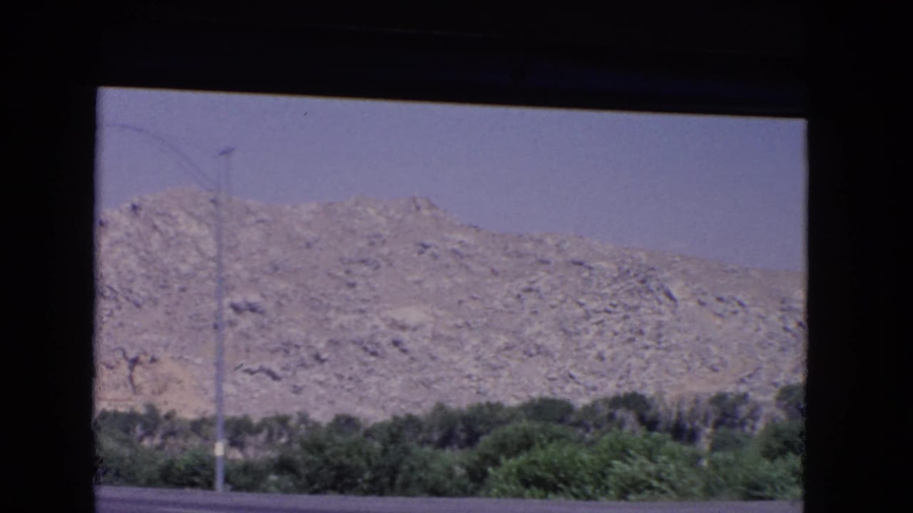 1966 California roadside landscape viewed through a car window, showing rocky hills, dark green shrubs, a tall pole, and the edge of the highway.