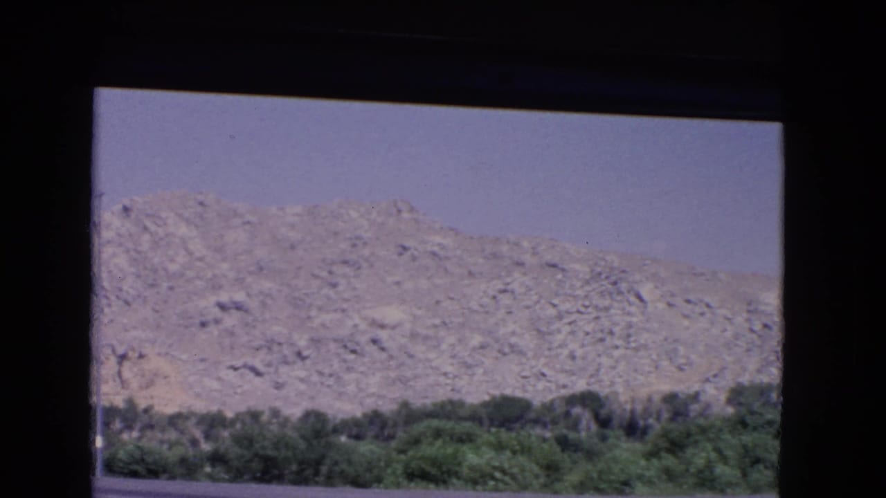 1966 California 8mm view of a rocky mountain range through the rear or side of a car, with green trees below a pale sky.