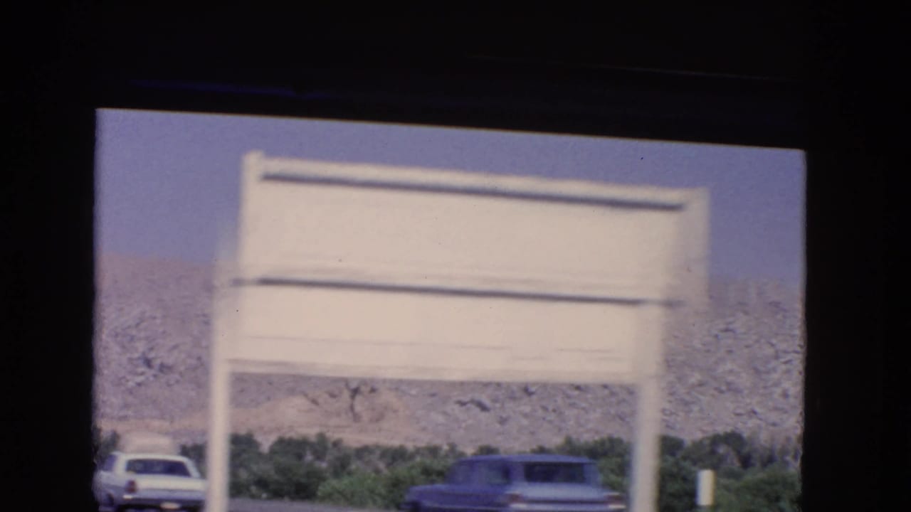 1966 California highway view with a blurred white roadside sign, two cars, trees, and a rocky hillside beyond.