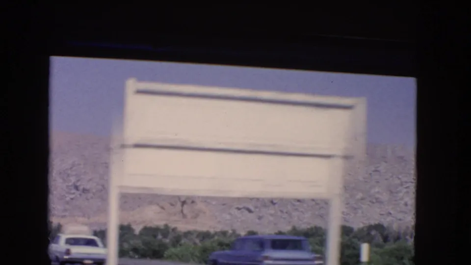 1966 California highway view with a blurred white roadside sign, two cars, trees, and a rocky hillside beyond.