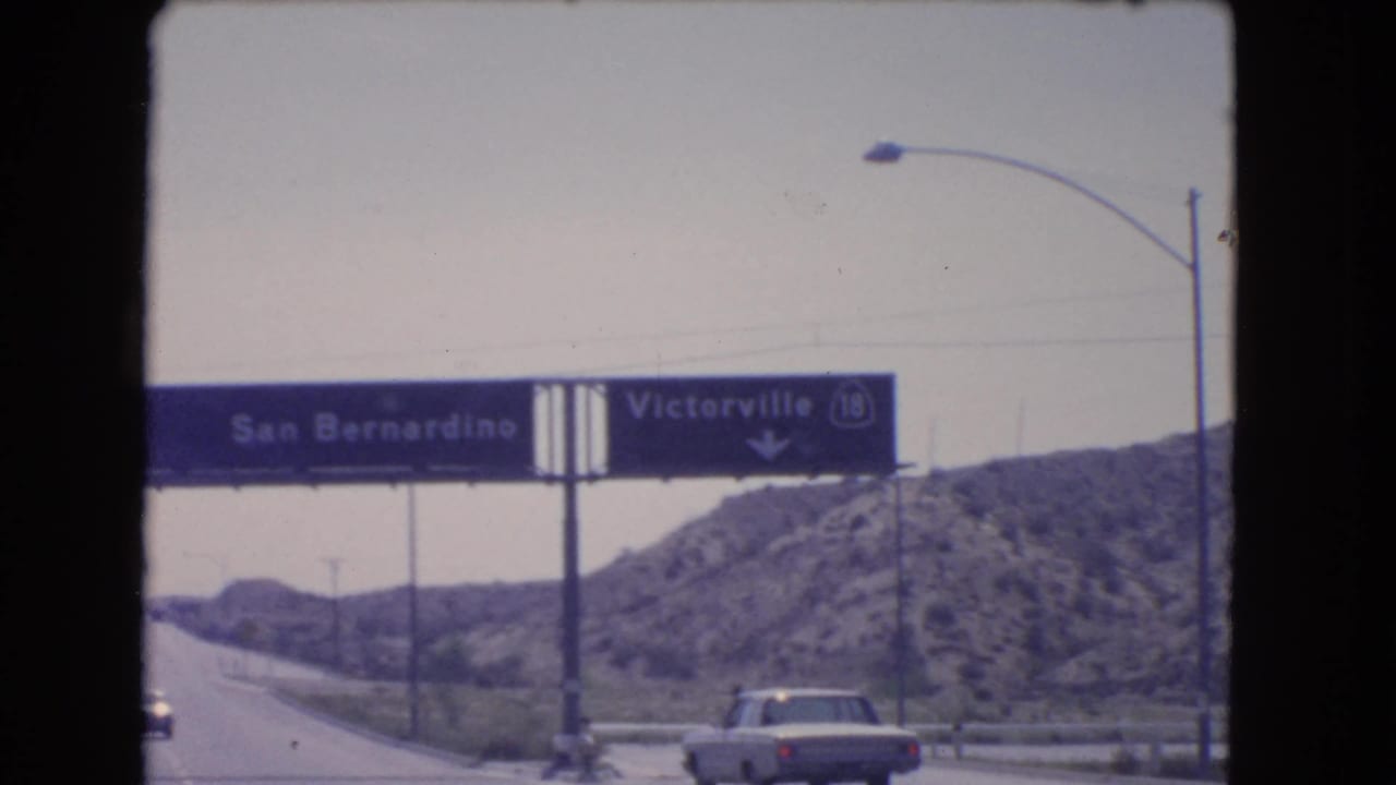 Overhead highway sign for San Bernardino and Victorville above cars near rocky hills.