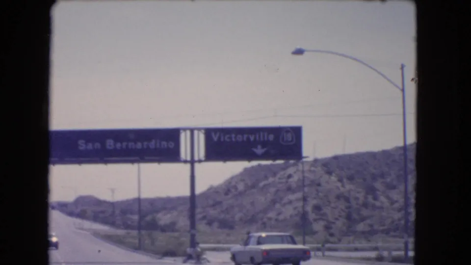 1966 8mm frame showing an overhead sign toward San Bernardino and Victorville on Highway 18, with cars and rocky hills nearby.
