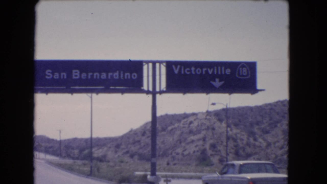 Close 1966 8mm view of a blue highway sign for San Bernardino and Victorville, with a car and rocky hillside below.