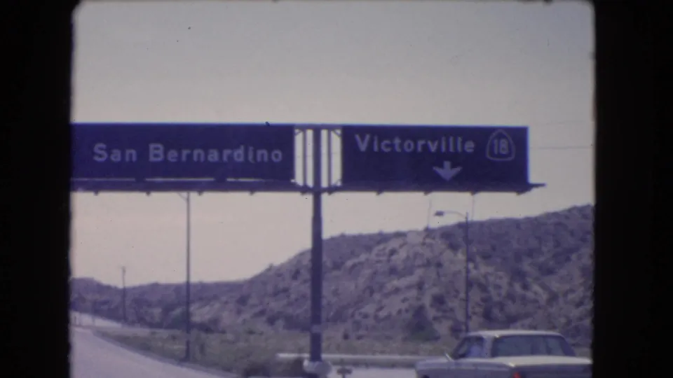 Close 1966 8mm view of a blue highway sign for San Bernardino and Victorville, with a car and rocky hillside below.