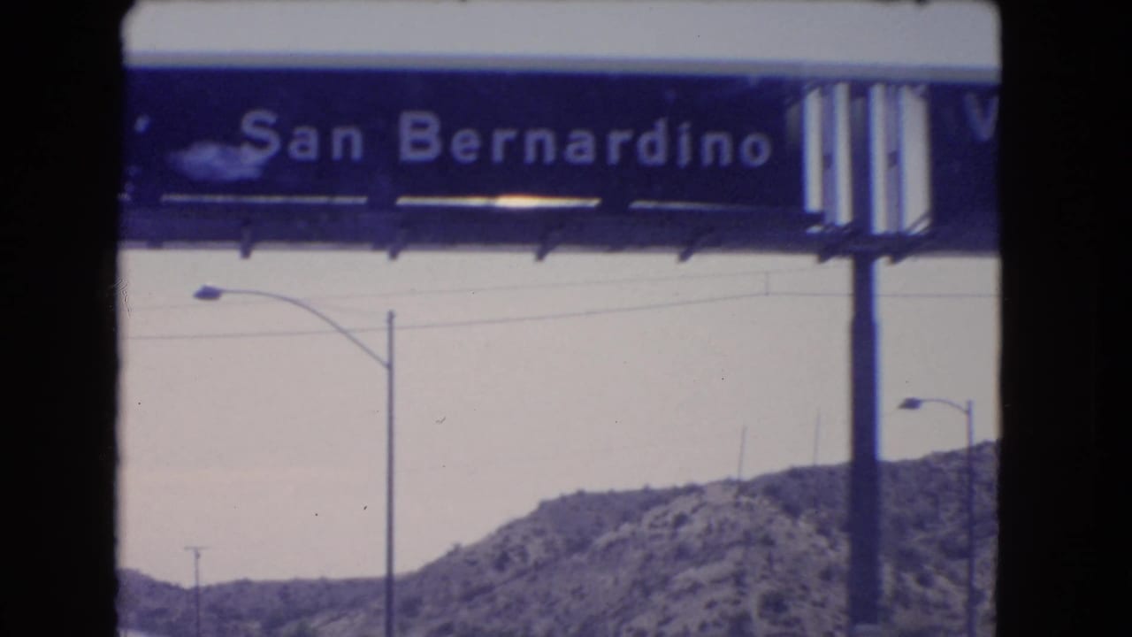 Soft 8mm close frame of a San Bernardino highway sign over a road corridor with streetlights and rocky Southern California hills.
