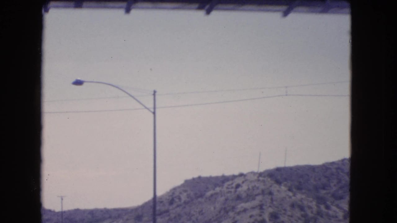 1966 California home-movie frame showing a roadside streetlamp, power lines, pale sky, and rocky hills from a moving car.