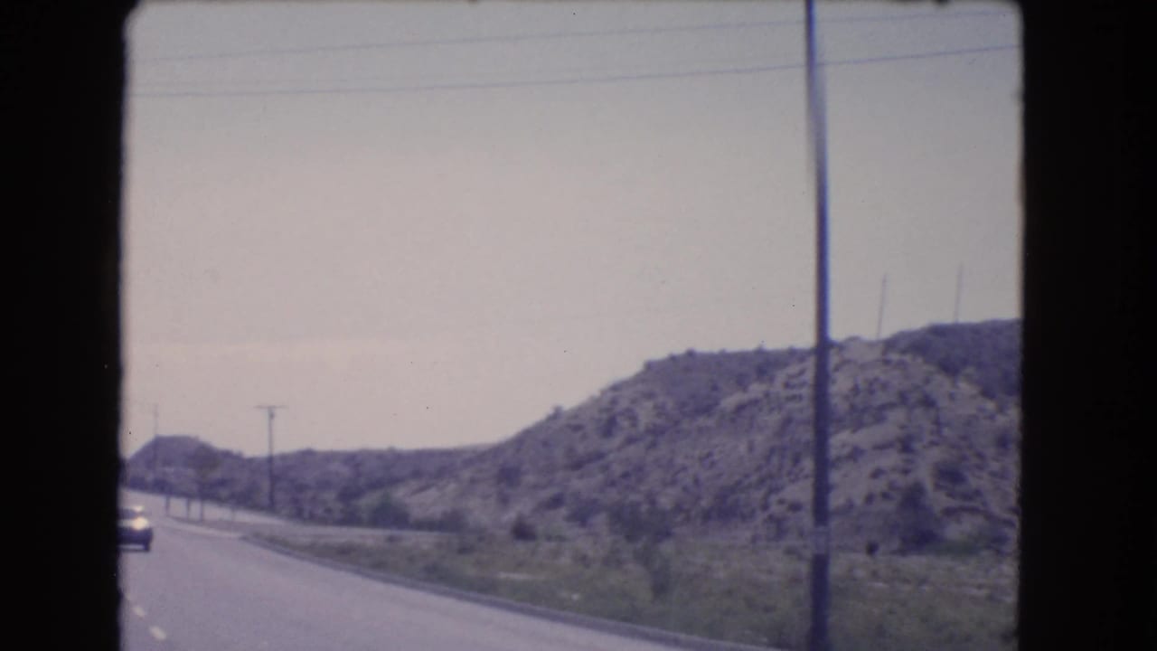 1966 California road-trip view of a highway curving along rocky hills, with utility poles and a distant vehicle visible ahead.