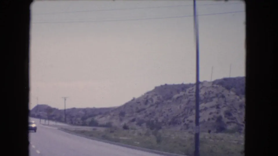 1966 California road-trip view of a highway curving along rocky hills, with utility poles and a distant vehicle visible ahead.