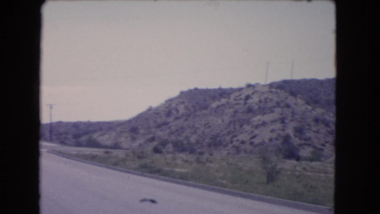 1966 California highway view from a moving vehicle, showing the road edge, roadside grass, utility poles, and a rocky hillside.