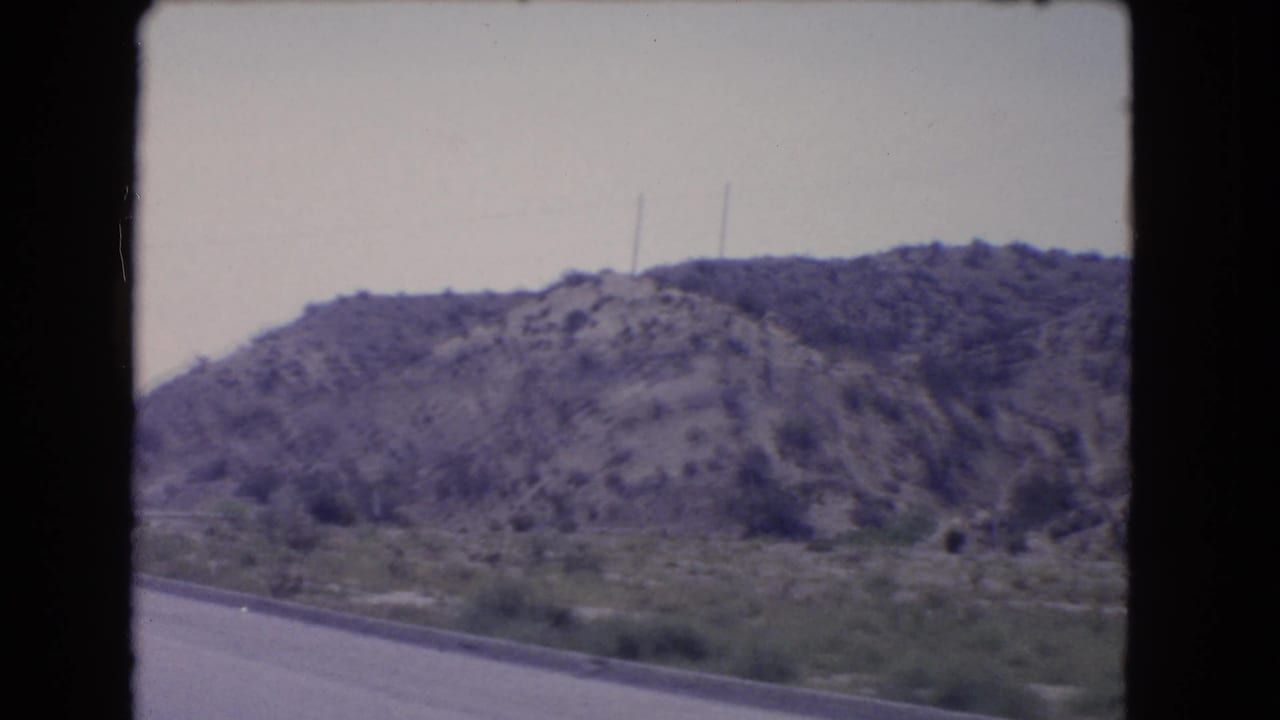 1966 California home-movie frame looking past a highway edge toward scrubby vegetation, utility poles, and rocky hills.