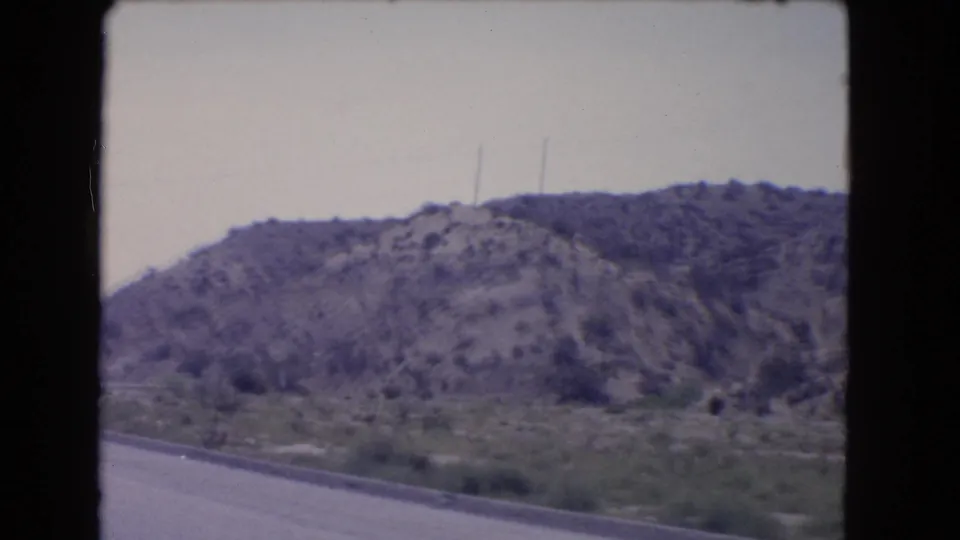 1966 California home-movie frame looking past a highway edge toward scrubby vegetation, utility poles, and rocky hills.