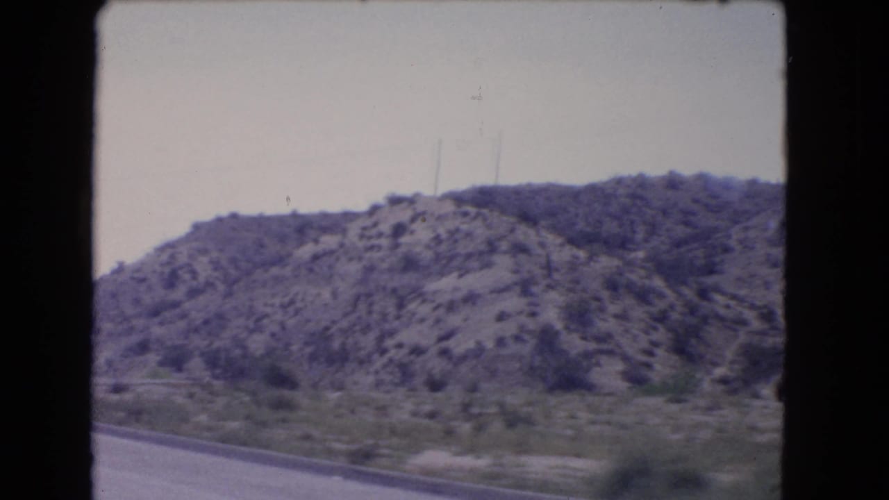 1966 California 8mm view of an arid roadside hillside with sparse shrubs and utility poles under a pale sky.