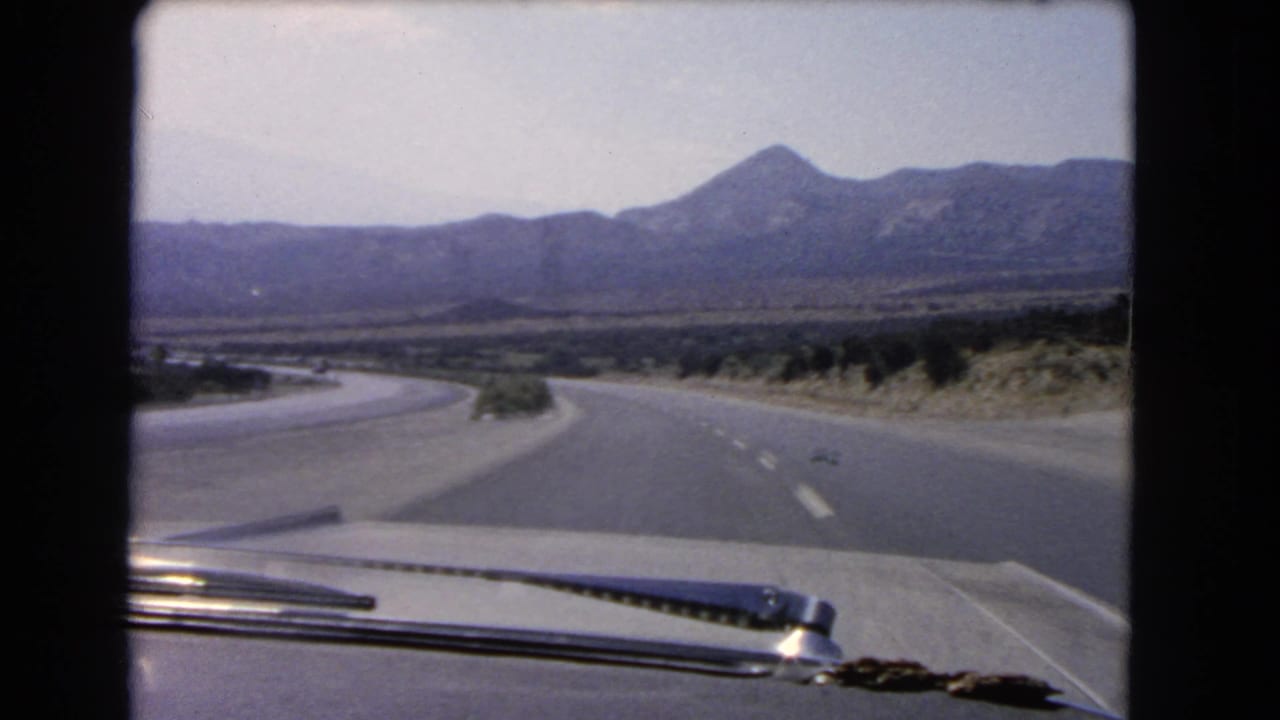 Faded 8mm windshield view from a moving car on a California desert highway with mountains on the horizon.
