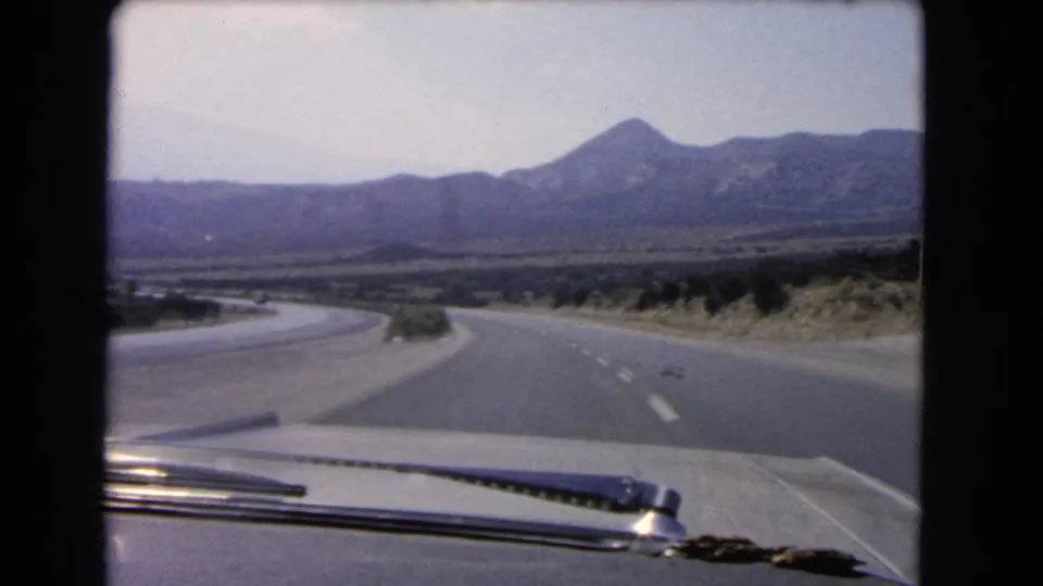 Faded 8mm windshield view from a moving car on a California desert highway with mountains on the horizon.