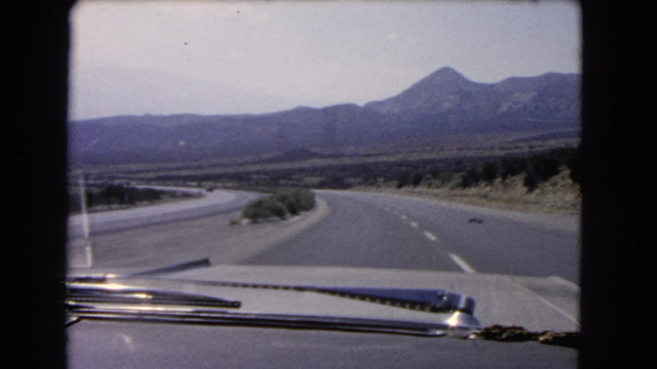 Passenger-side 8mm view of a California desert highway curve, with the car hood and windshield wiper framing the mountains ahead.