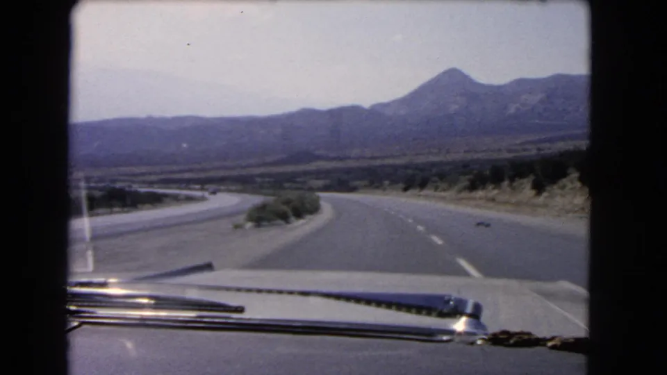 Passenger-side 8mm view of a California desert highway curve, with the car hood and windshield wiper framing the mountains ahead.