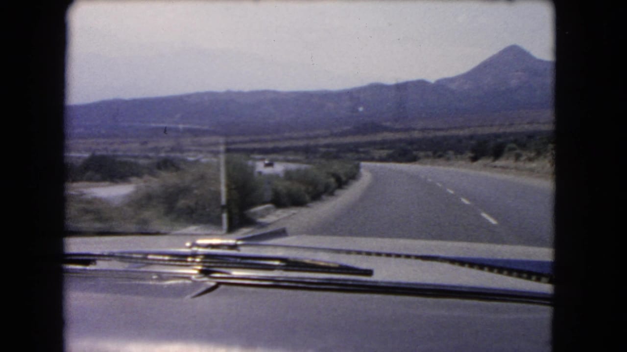 1966 California home-movie frame looking over a car hood at a curving highway, roadside shrubs, and mountains in the distance.