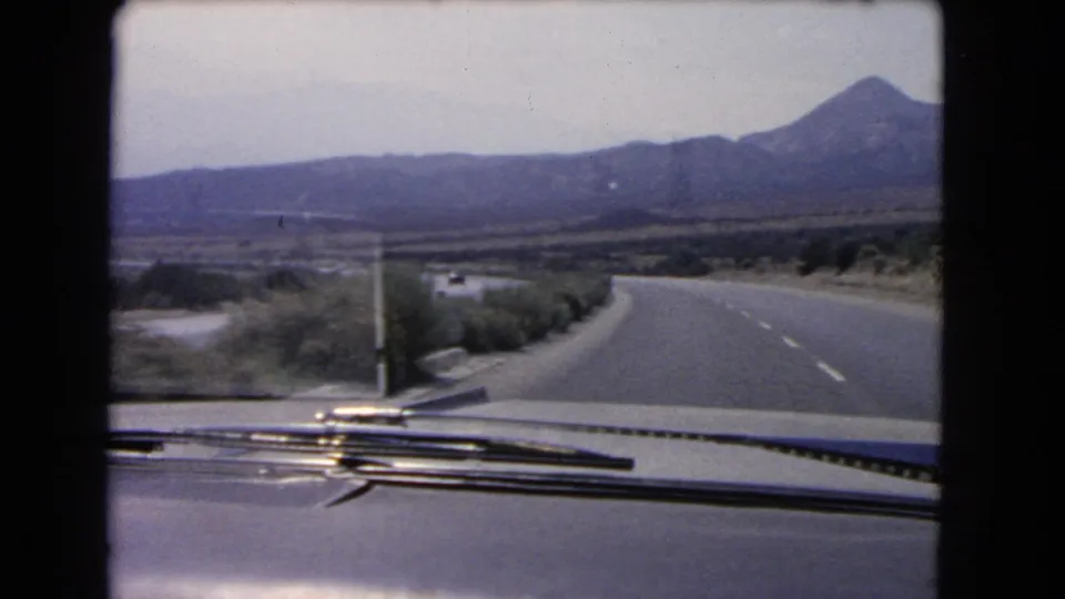 1966 California home-movie frame looking over a car hood at a curving highway, roadside shrubs, and mountains in the distance.