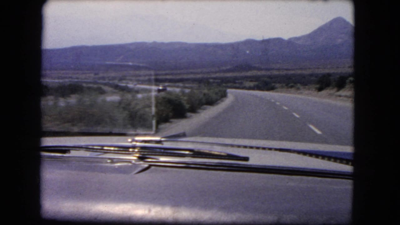 8mm frame from a 1966 California road trip showing the car hood, wipers, desert brush, and a highway leading toward mountains.