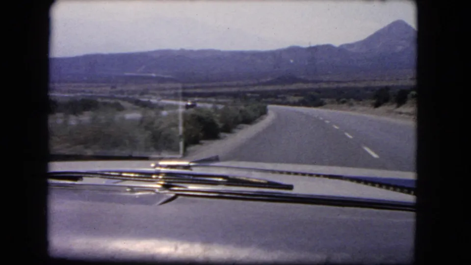 8mm frame from a 1966 California road trip showing the car hood, wipers, desert brush, and a highway leading toward mountains.