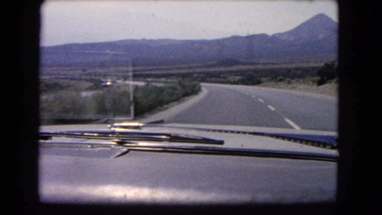 1966 California 8mm car-view frame with windshield wipers, a curving highway, low bushes, valley floor, and mountains ahead.