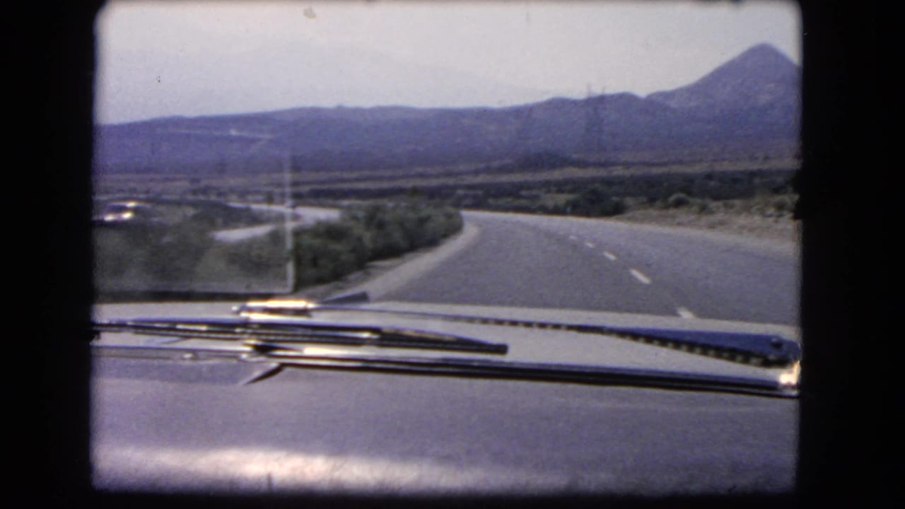 1966 California home-movie view through a car windshield toward a quiet curving highway, shrubs, and mountains.
