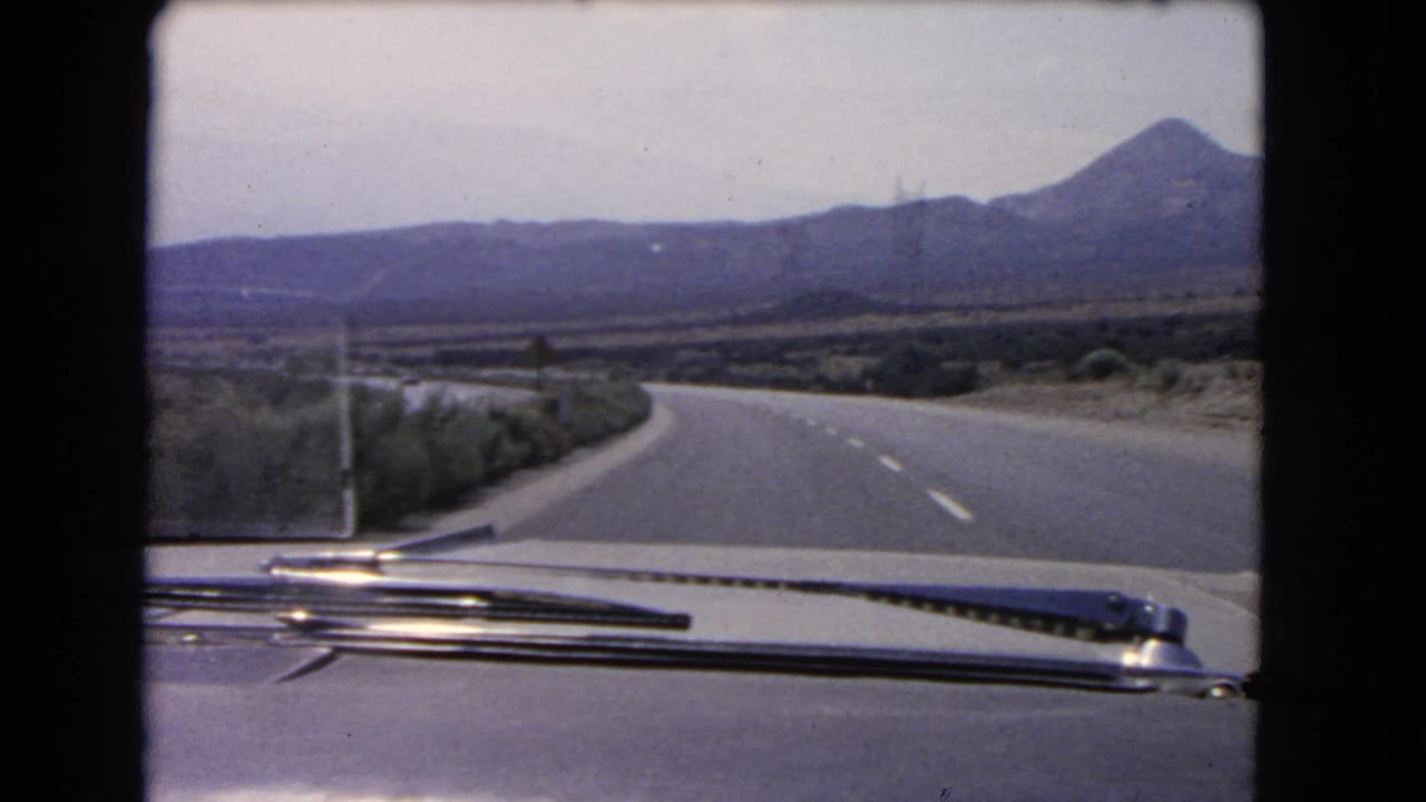 A 1966 California road-trip frame shows an empty two-lane highway curving across open scrubland below distant hills.