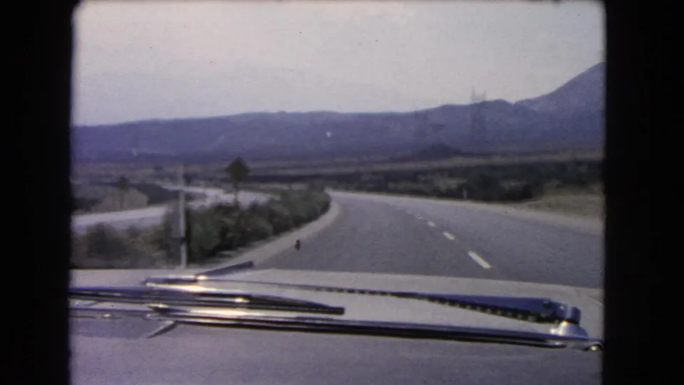 From inside the car, the road curves past a small sign in a hazy California mountain landscape from 1966.