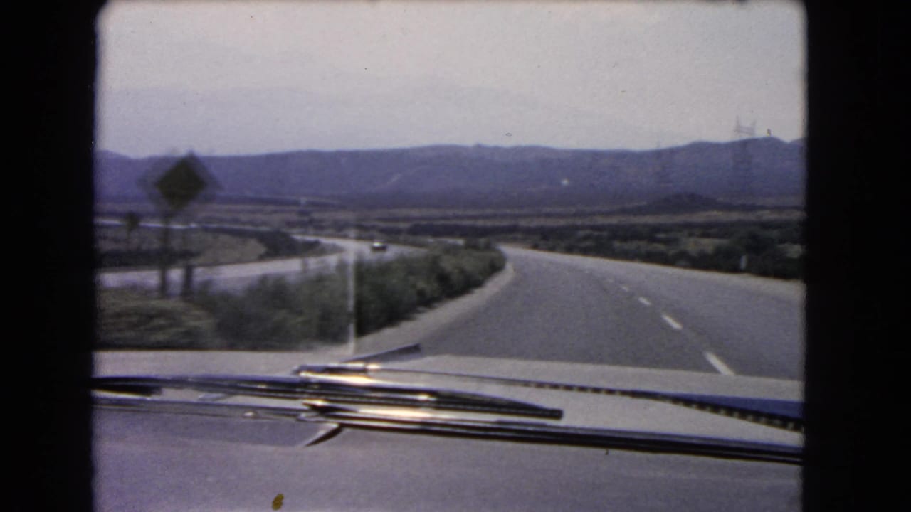 The final 1966 California frame looks over the car hood to a quiet highway, a roadside sign, sparse shrubs, and hazy hills.