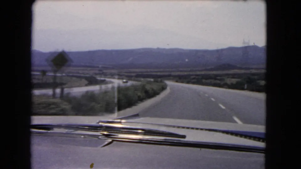 The final 1966 California frame looks over the car hood to a quiet highway, a roadside sign, sparse shrubs, and hazy hills.