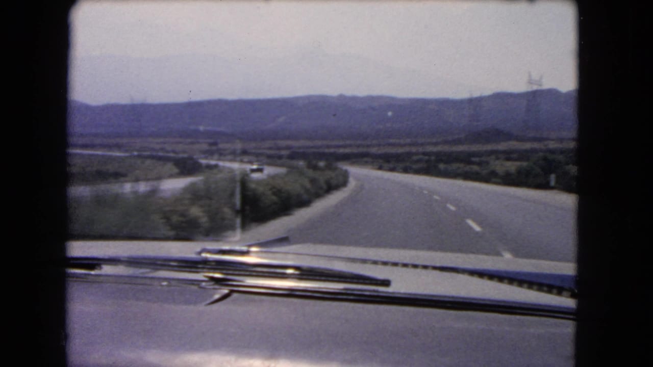 Car hood and wipers seen from inside a vehicle on a curving highway with hills ahead.