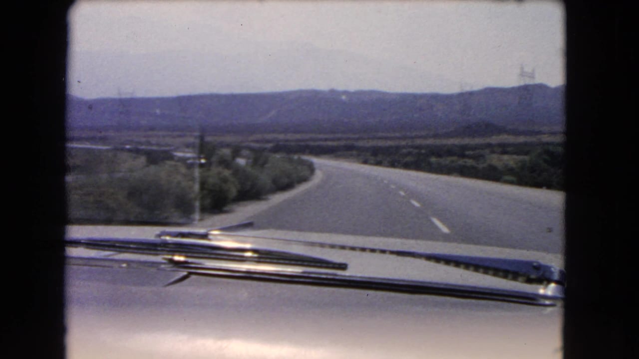 Forward road view from a moving car with dashboard details and low hills in the distance.