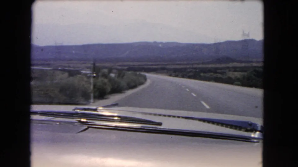 A car-mounted 8mm view looks down an open California highway, with windshield wipers in front and hazy hills beyond.
