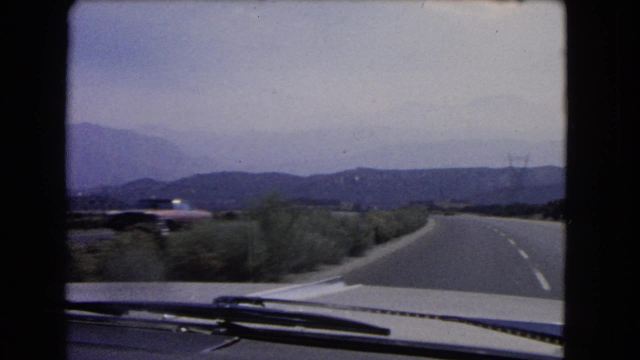 1960s home-movie footage from inside a car shows a right-bending California highway bordered by brush and distant mountains.