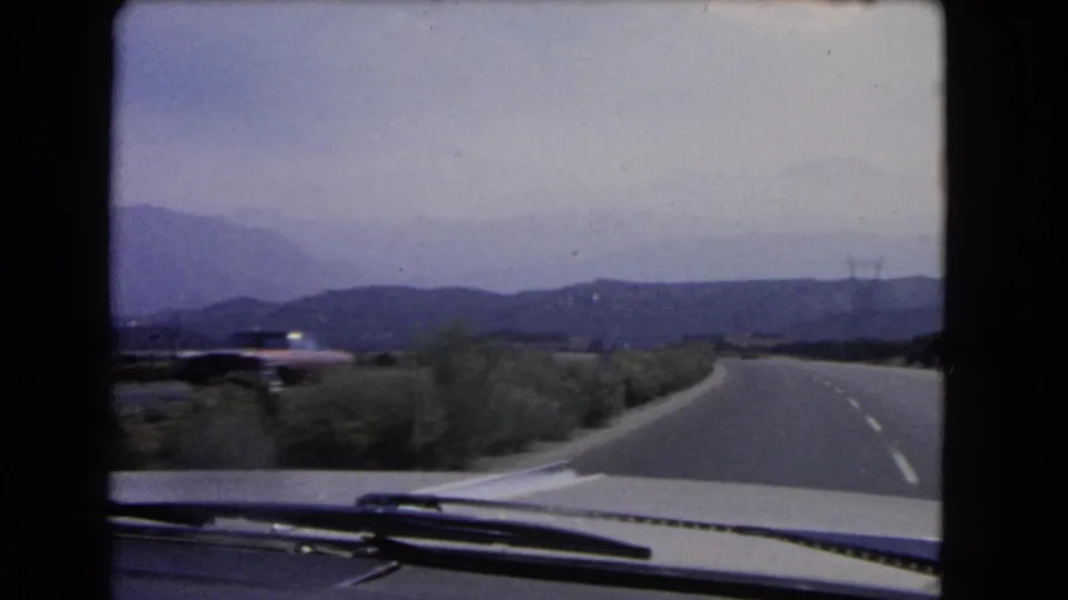 1960s home-movie footage from inside a car shows a right-bending California highway bordered by brush and distant mountains.