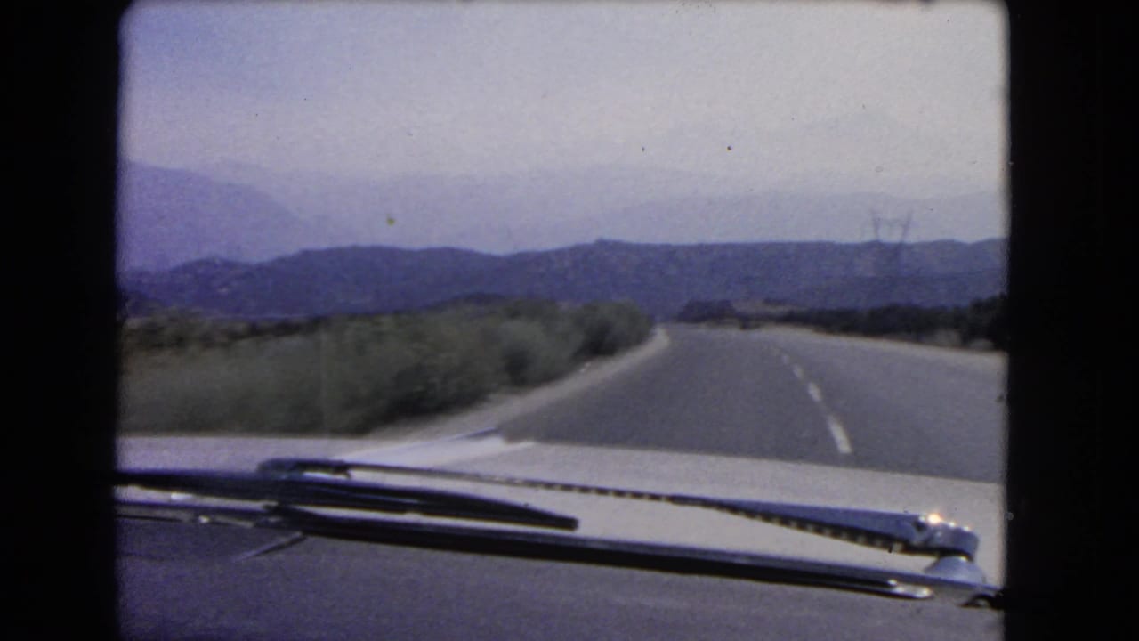 1966 California 8mm road-trip frame showing a car-level view of a paved mountain road, distant hills, and windshield hardware.