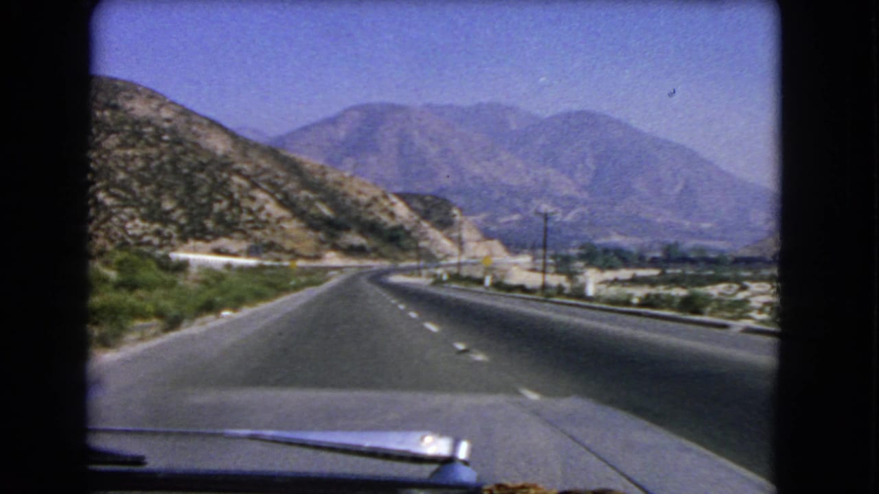 1966 California home-movie frame showing a forward car view of a curving mountain road with scrub vegetation and distant peaks.