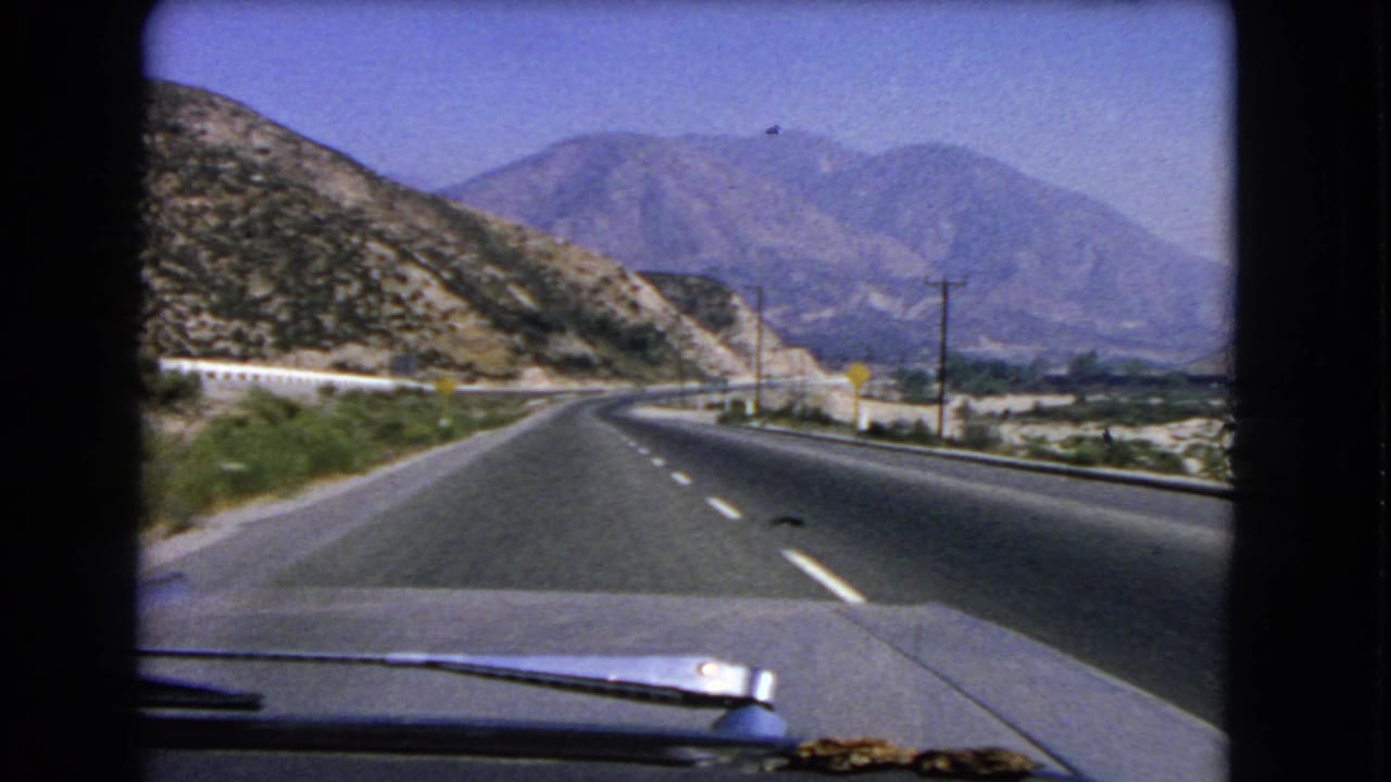 1966 California 8mm frame of a road-trip view, with the car hood below a winding mountain roadway, poles, slopes, and distant hills.