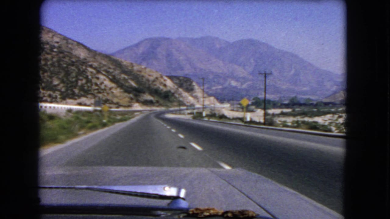 1966 California home-movie frame from inside a car, showing a curving highway, roadside signs, dry hills, and mountains ahead.