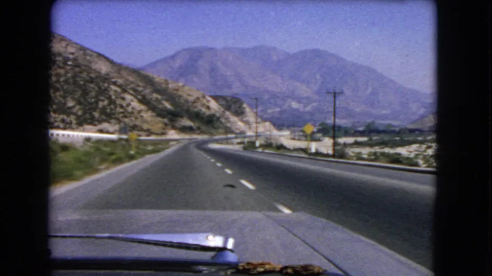 1966 California home-movie frame from inside a car, showing a curving highway, roadside signs, dry hills, and mountains ahead.