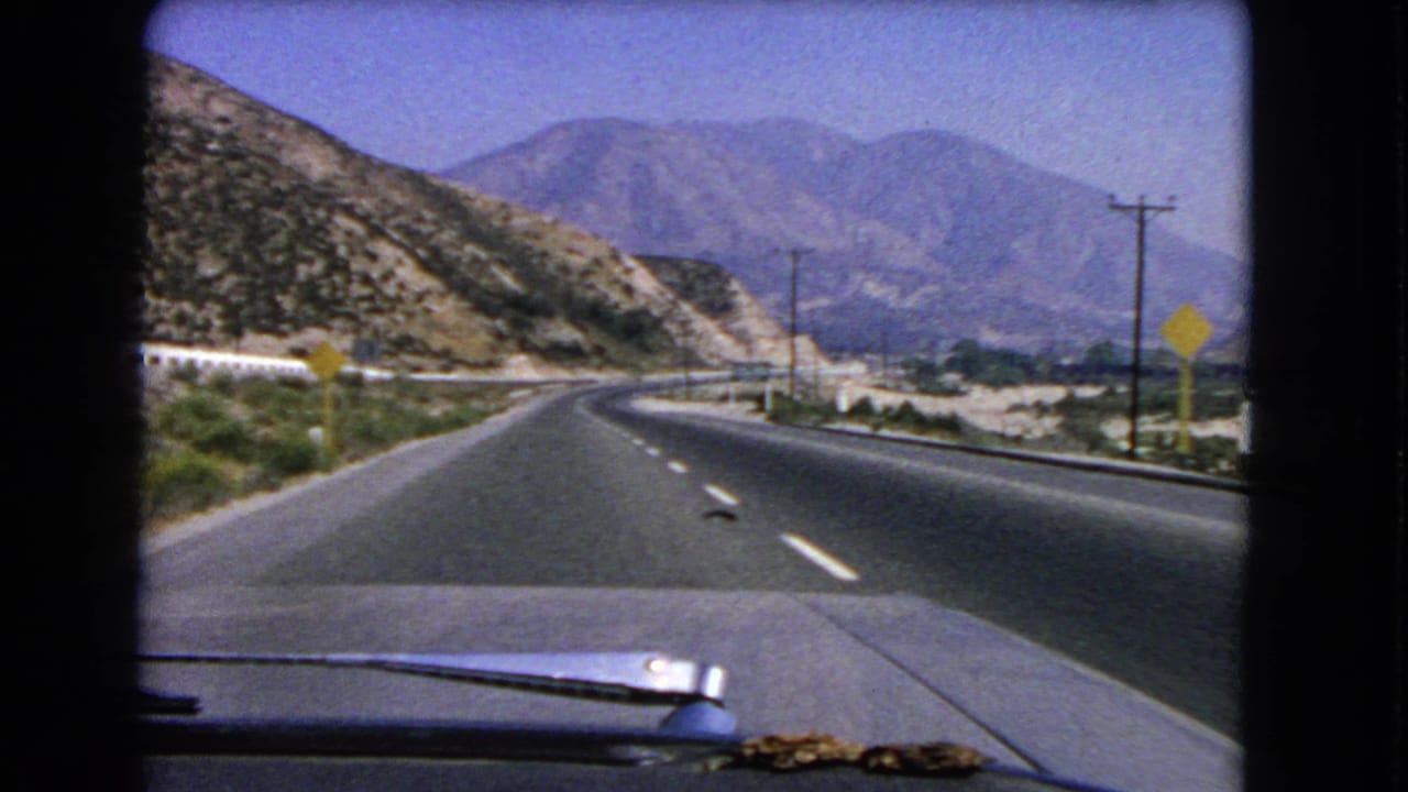 Inside-car view on a 1966 California highway, with the road bending through arid hills and mountains under a clear sky.