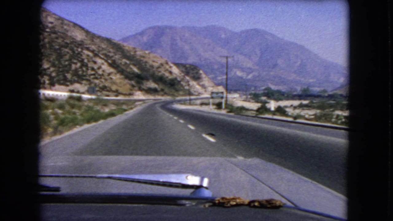 1966 California windshield view showing a two-lane highway, rocky slope, utility poles, and distant mountain ridges.