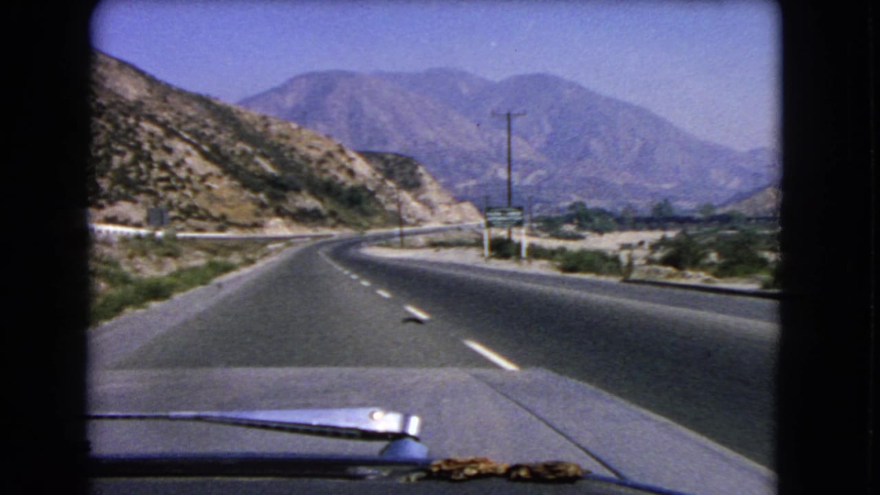 Final frame of a 1966 California car drive, showing the highway curving through a dry valley with utility poles and mountains beyond.