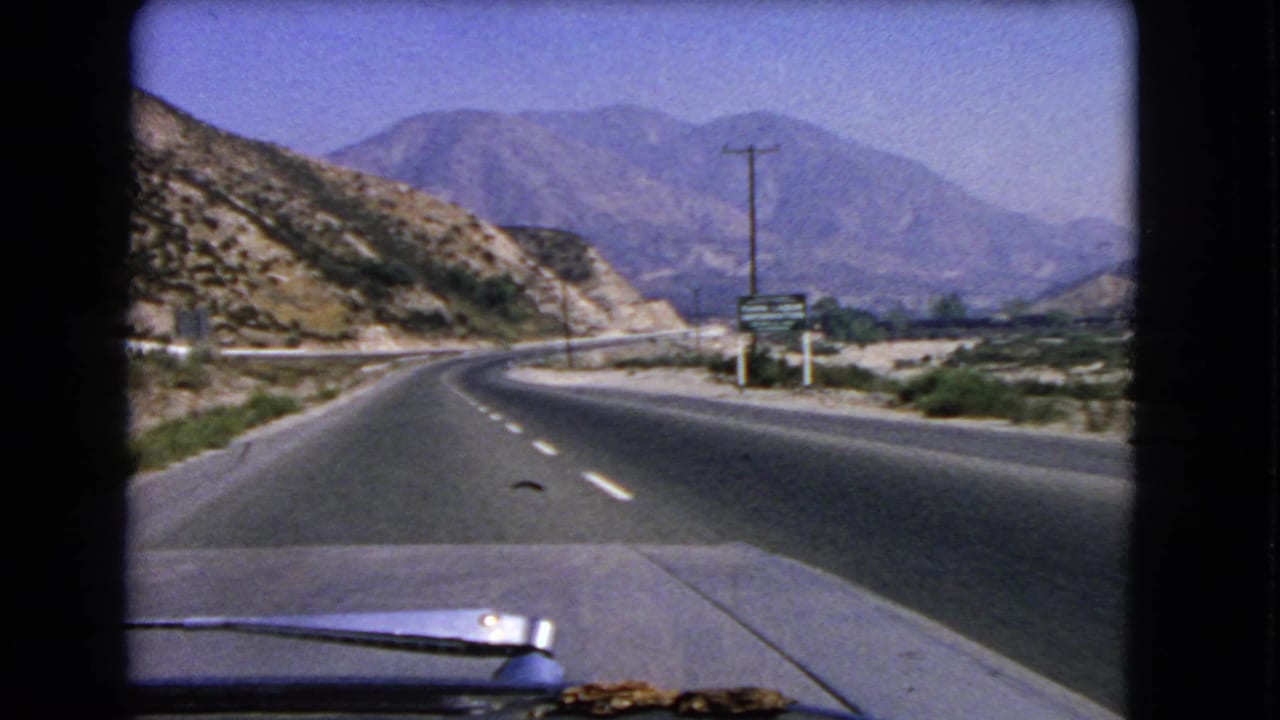 1966 California 8mm driving view along a curving mountain highway with a car hood in the foreground and dry hills beyond.