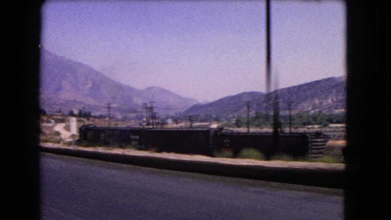 1966 California roadside view from a moving car, showing dark freight rail cars, utility poles, dry hills, and mountains.